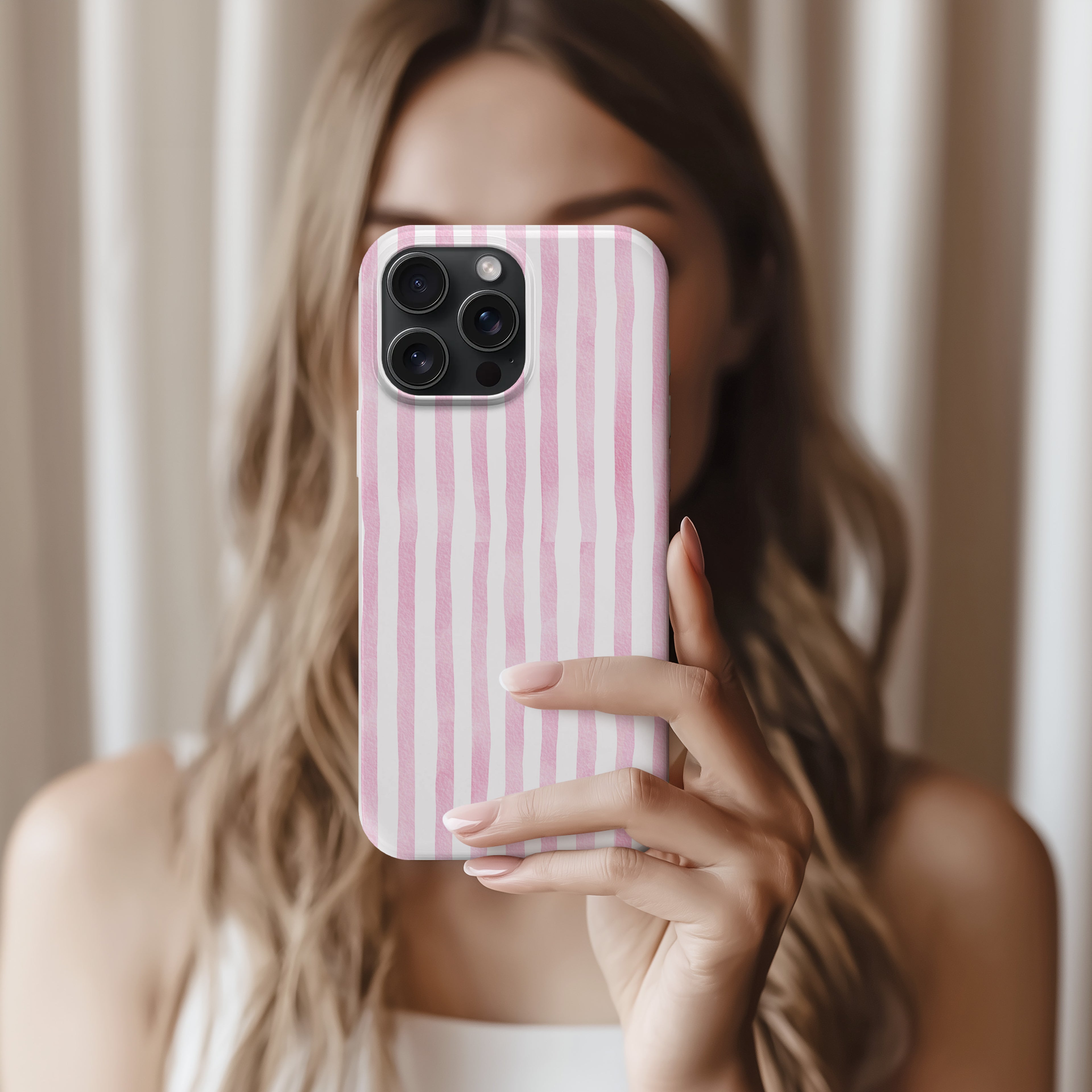 A woman with long, wavy hair is holding a pink and white striped phone case and taking a selfie.