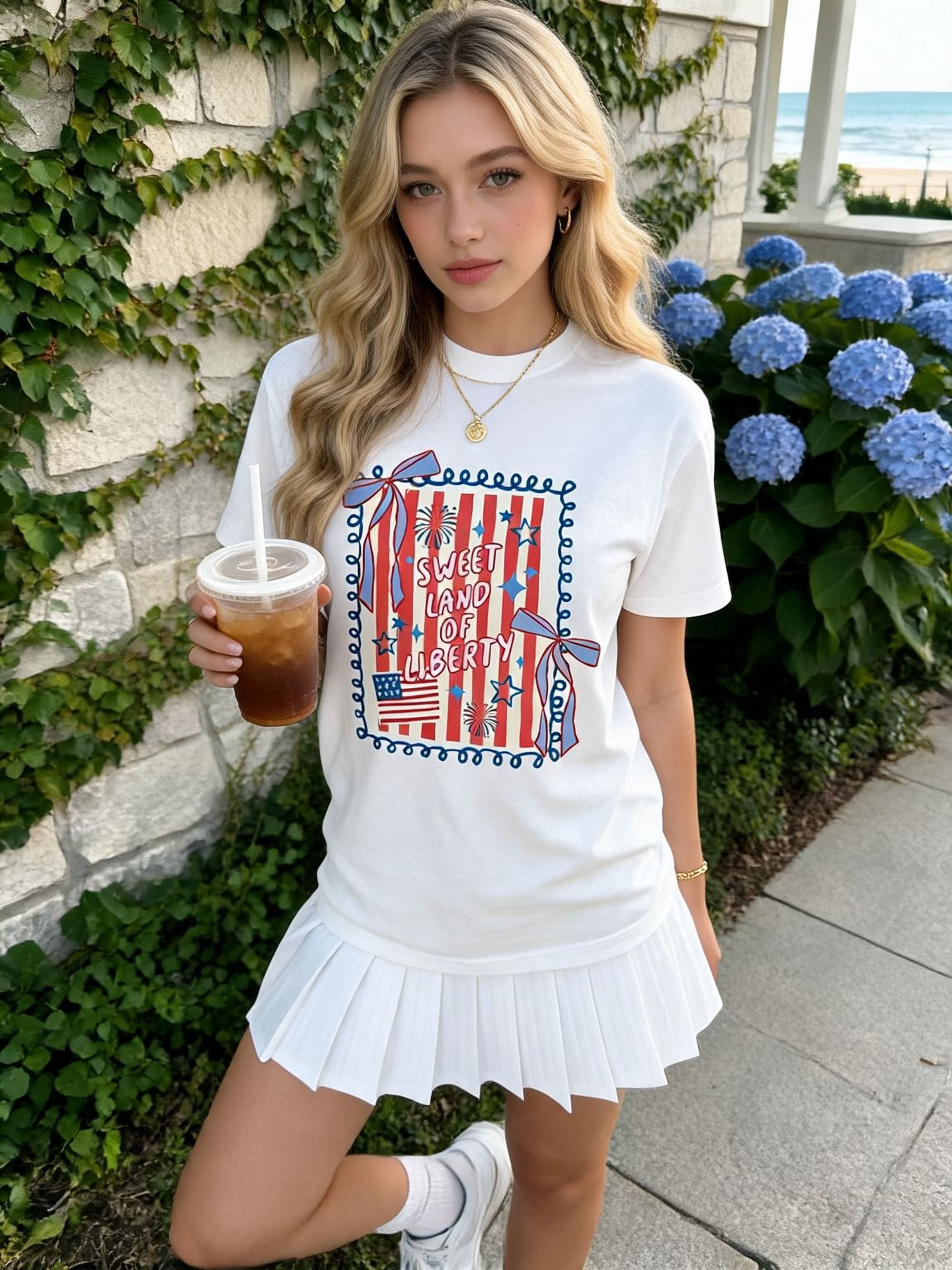 A young woman wearing a white skirt and a white t-shirt with a patriotic design is holding a drink and standing in front of a stone wall with blue flowers.