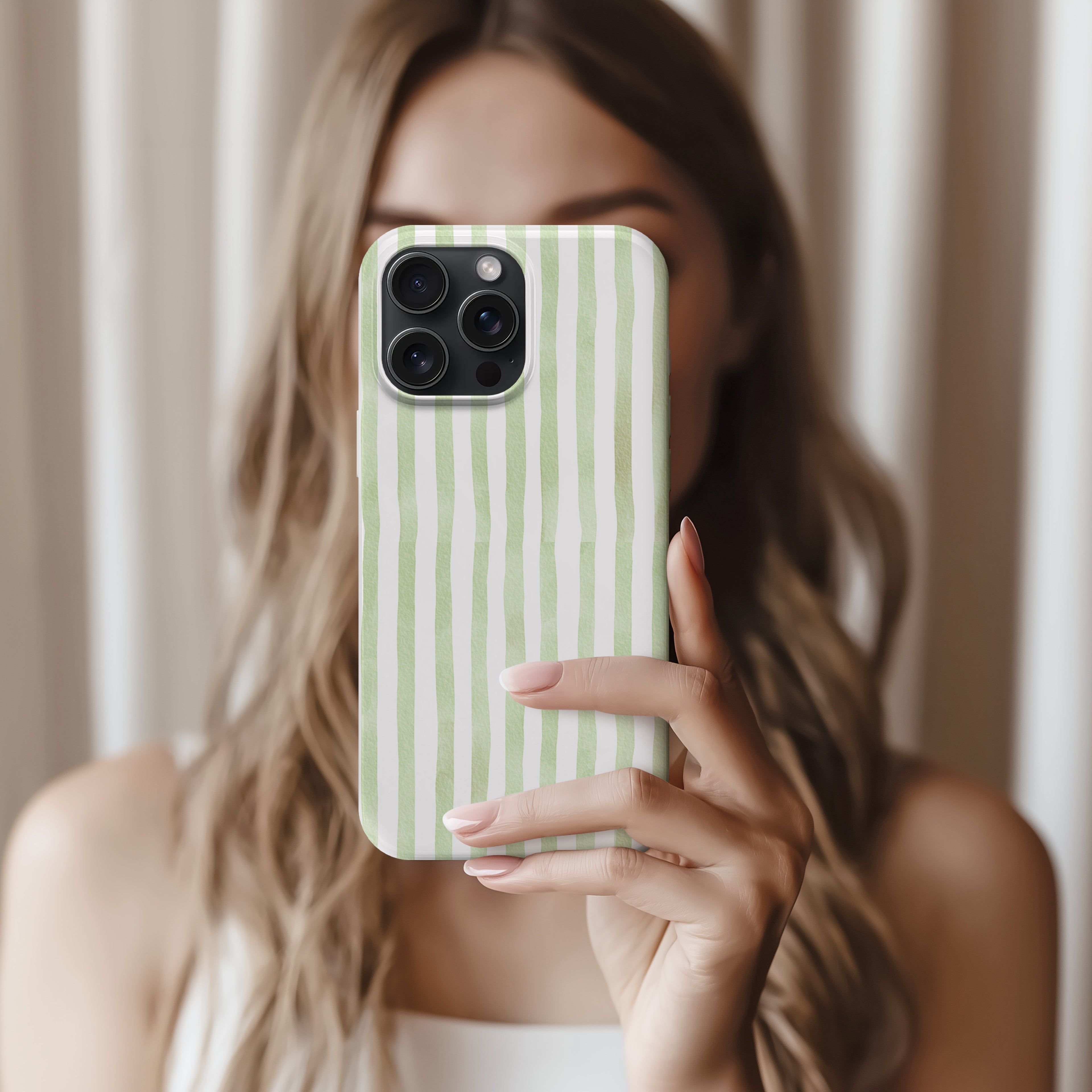A woman with long, wavy blonde hair is holding a phone with a green and white striped pattern, taking a selfie in front of a white curtain.