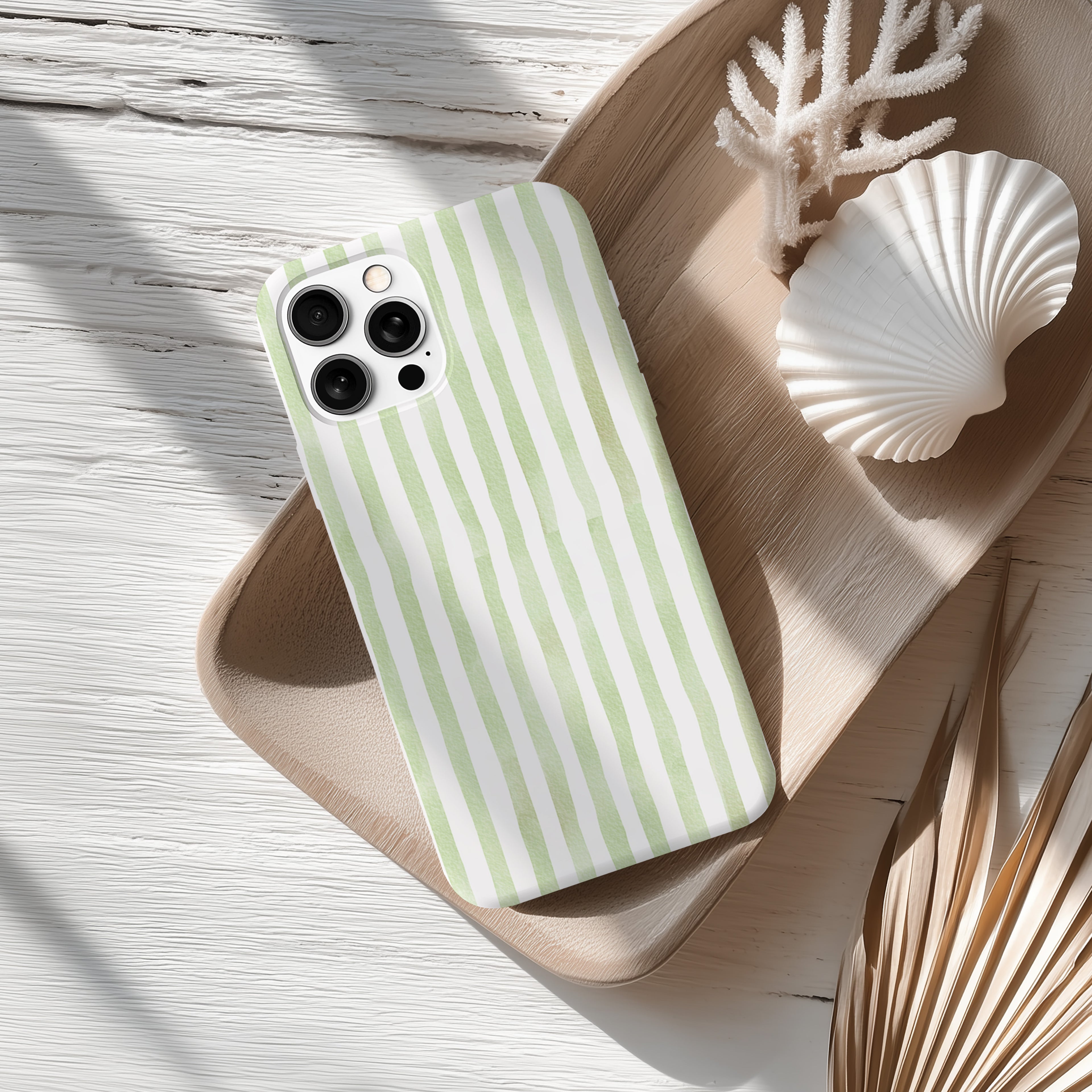 A green and white striped phone case is placed on a wooden surface, accompanied by a seashell and some dried leaves.