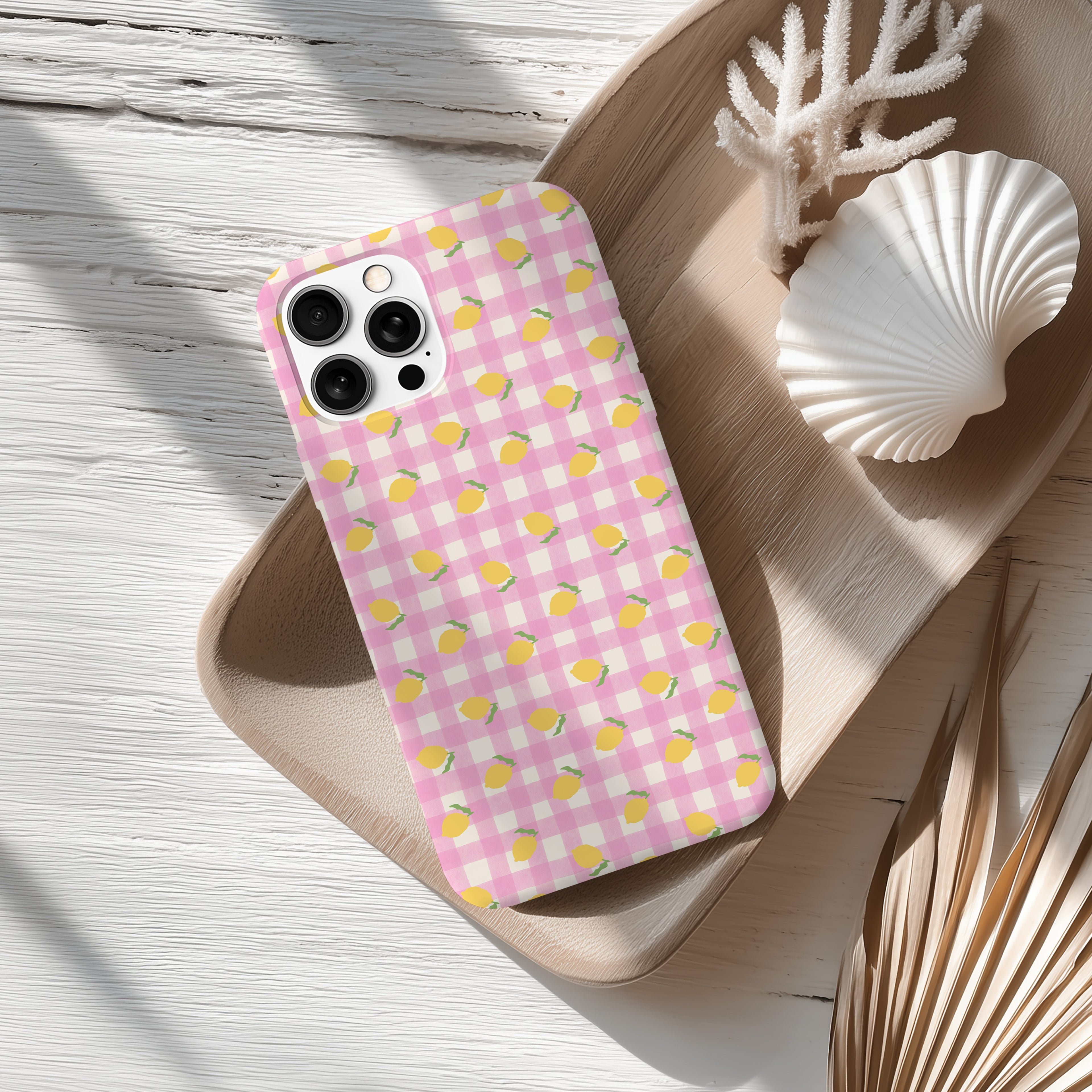 A pink and white checkered patterned phone case is placed on a wooden surface, accompanied by a white seashell and some dried leaves.