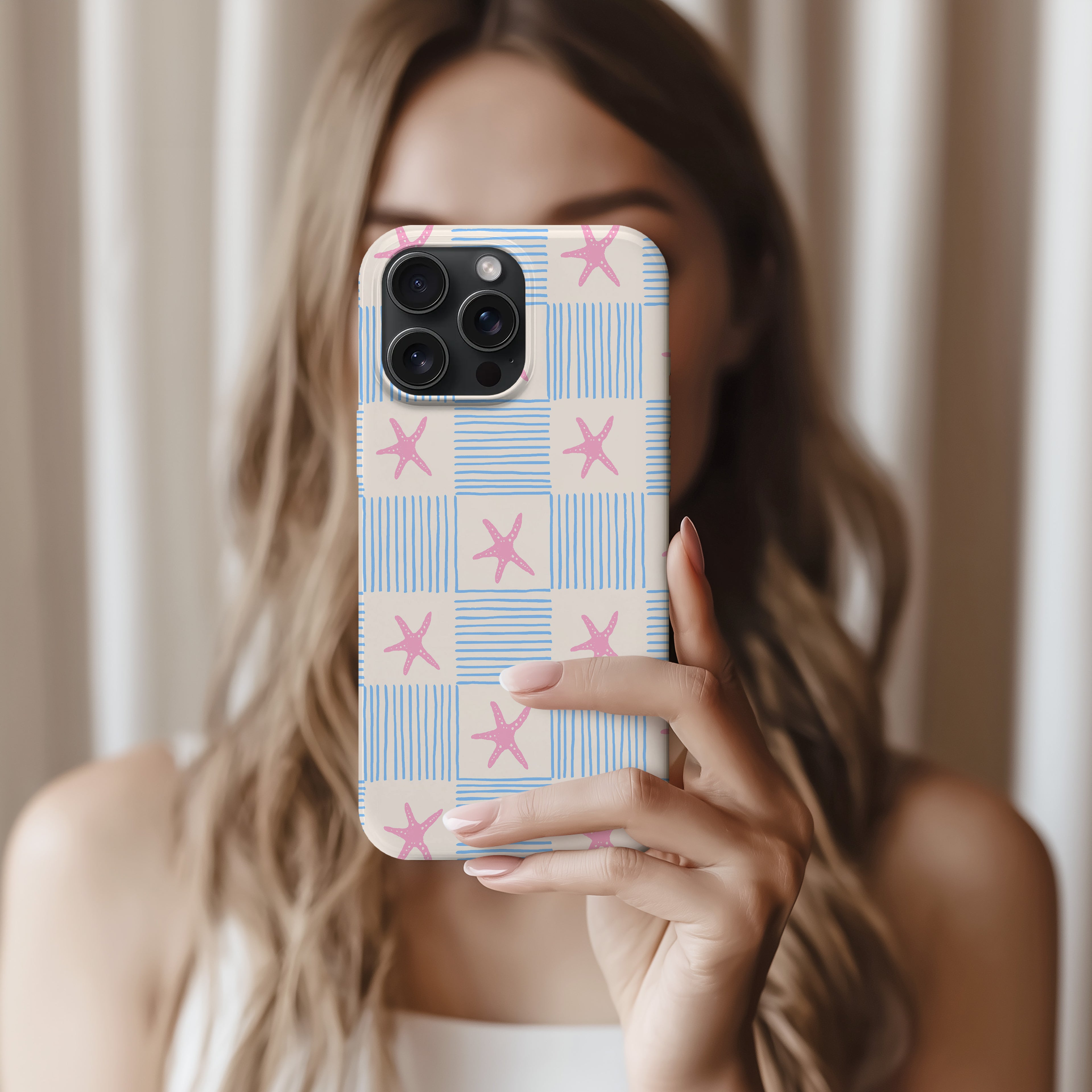 A woman is holding a smartphone with a patterned case, taking a selfie in front of a curtain.