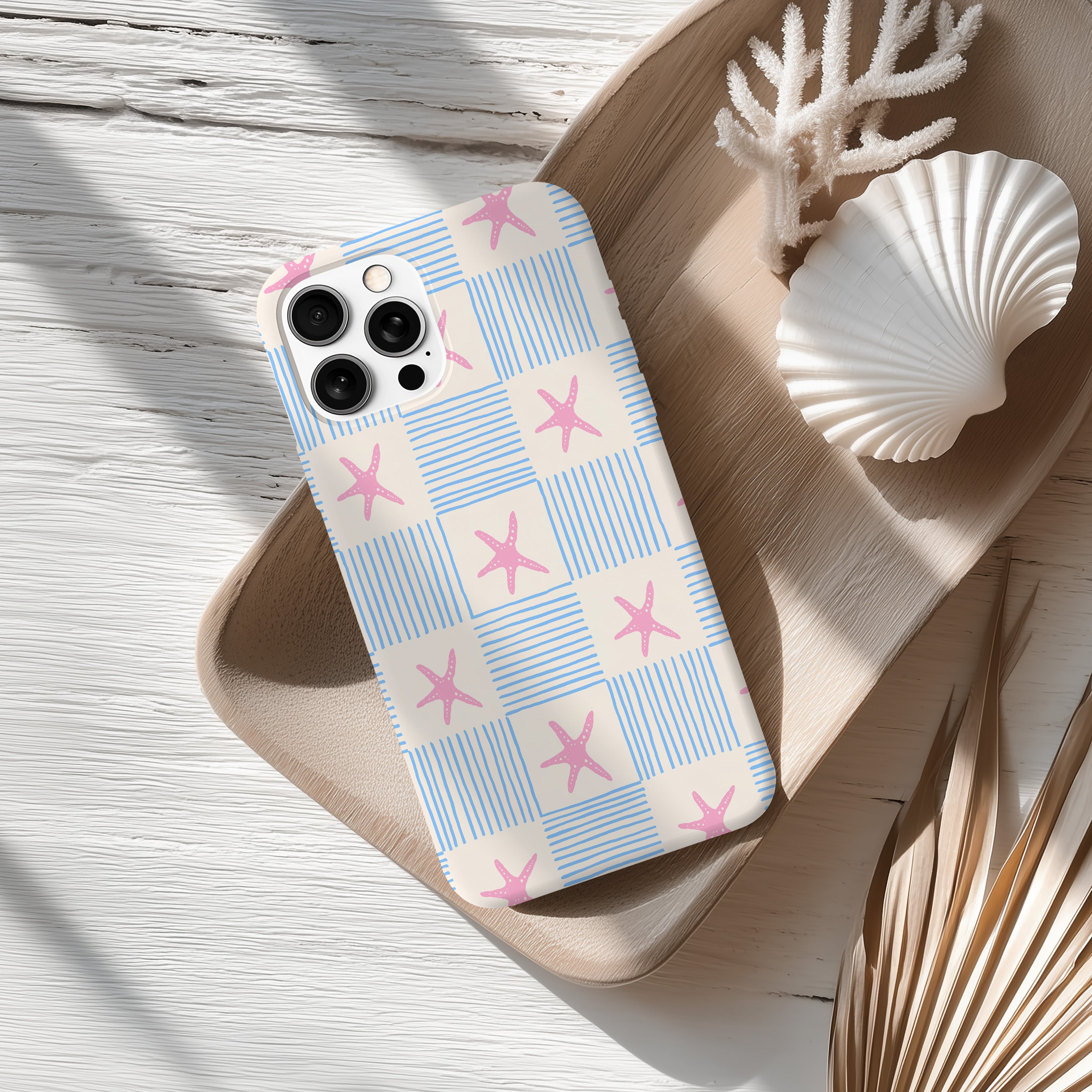 a smartphone case with a blue and white checkered pattern and pink star shapes, placed on a wooden surface alongside a seashell and some dried leaves.