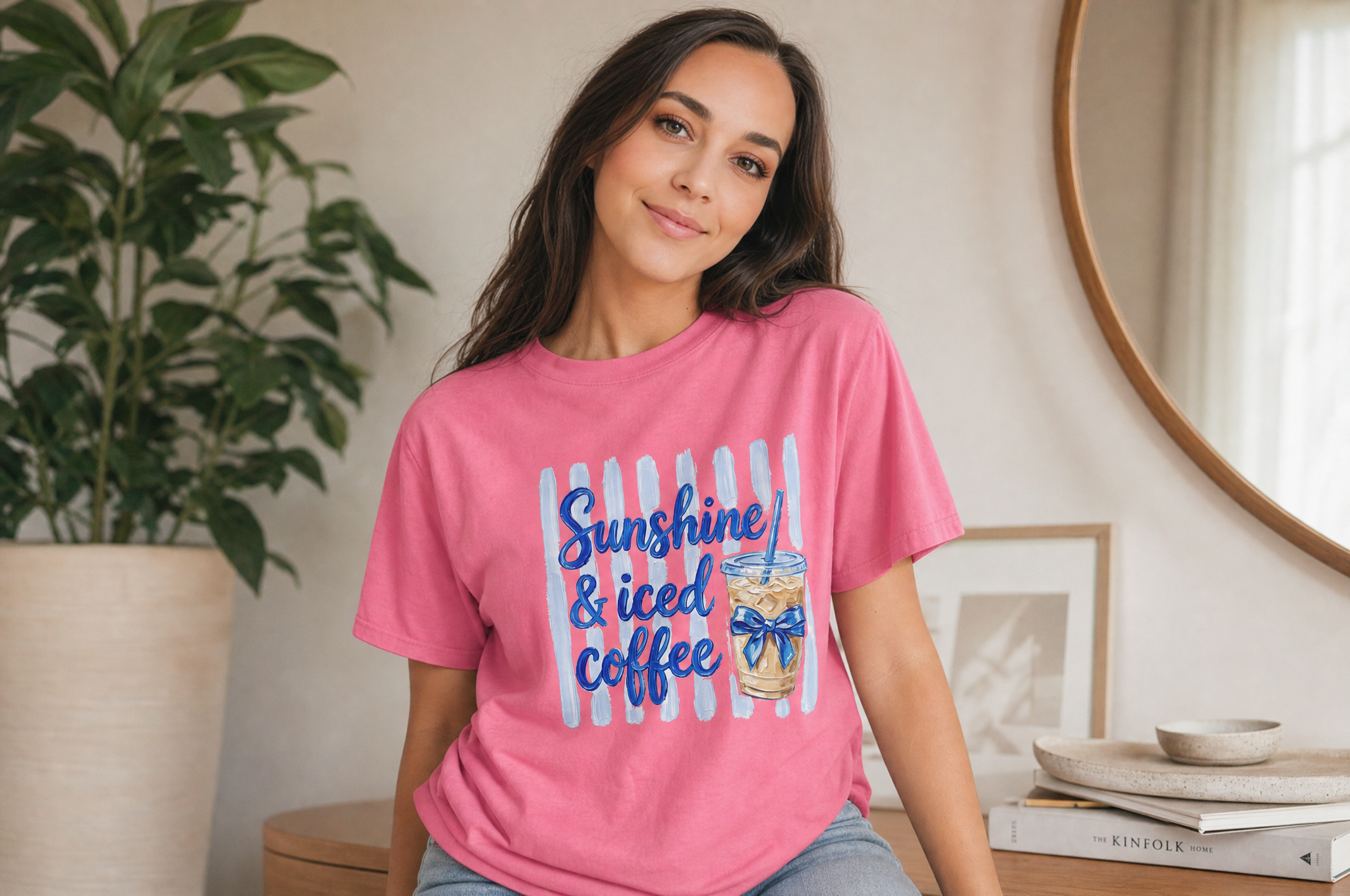 A young woman wearing a pink t-shirt with a graphic design of a glass of iced coffee and the text "Sunshine & iced coffee" is sitting in a room with a potted plant and a round mirror.
