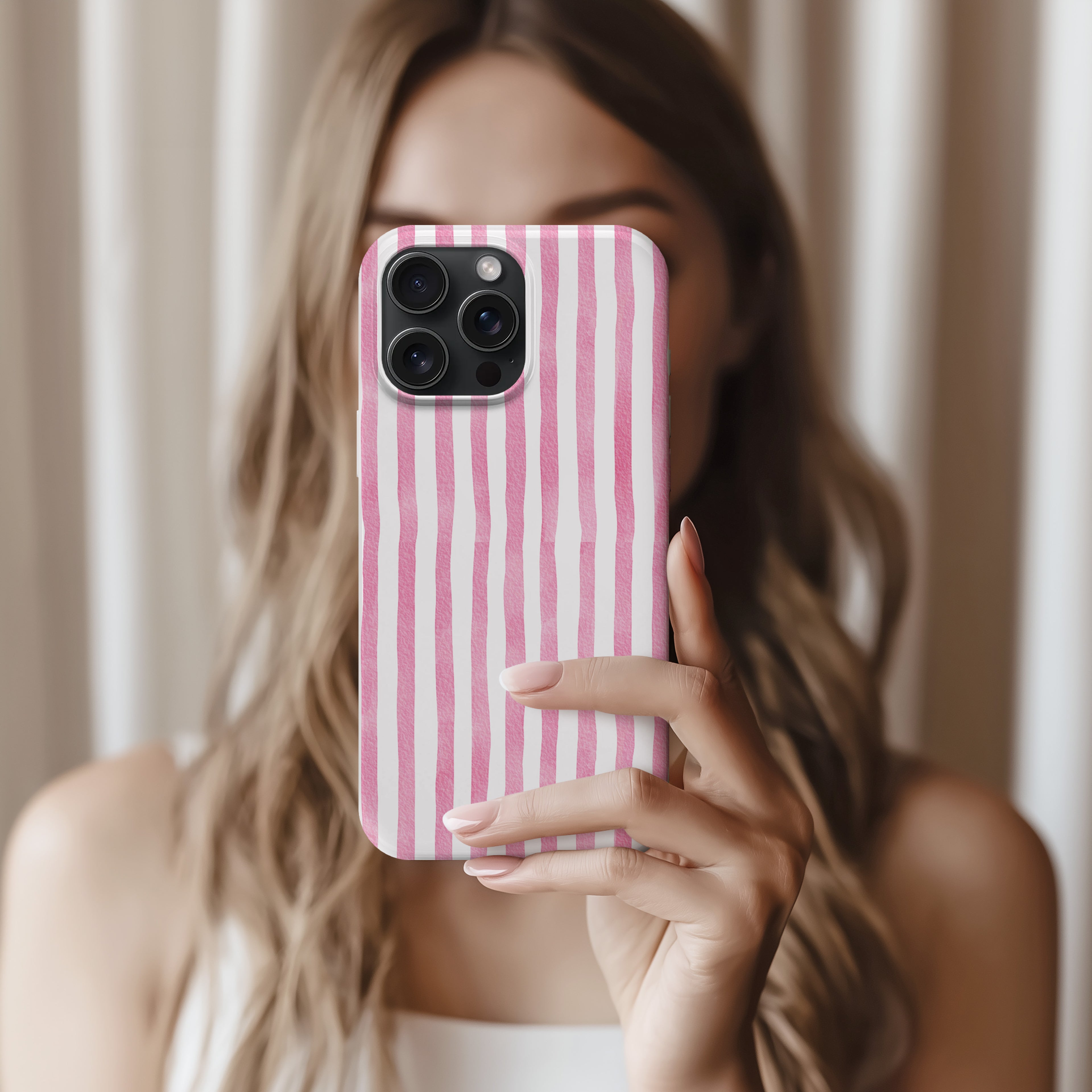 A woman with long, wavy hair is holding a pink and white striped phone case up to the camera, taking a selfie.